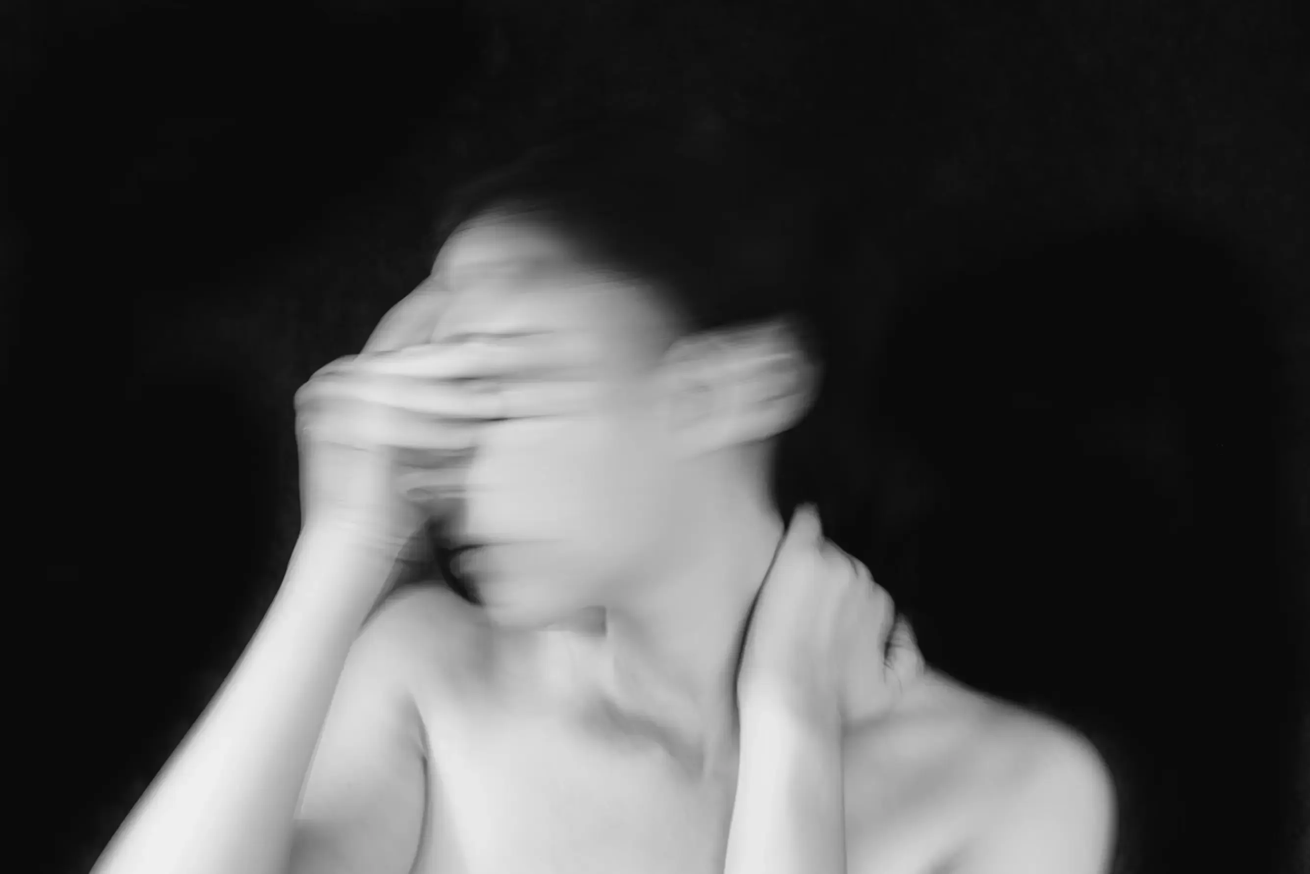 Black and white portrait, blurred by a long exposure, of a woman against a black background: one hand covers her eyes and the other holds the back of her neck.