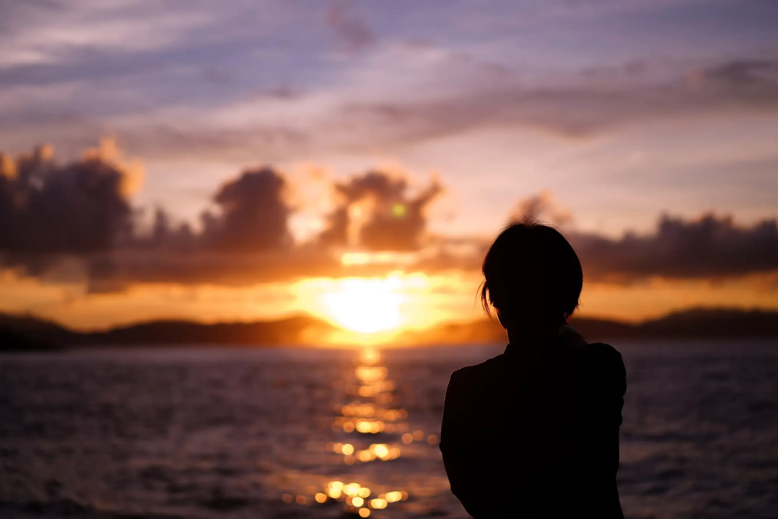 Silhouette in controluce di una persona di spalle, in primo piano a destra, che guarda il sole tramontare sul mare fra nuvole arancioni; la luce traccia una scia dorata sull'acqua.