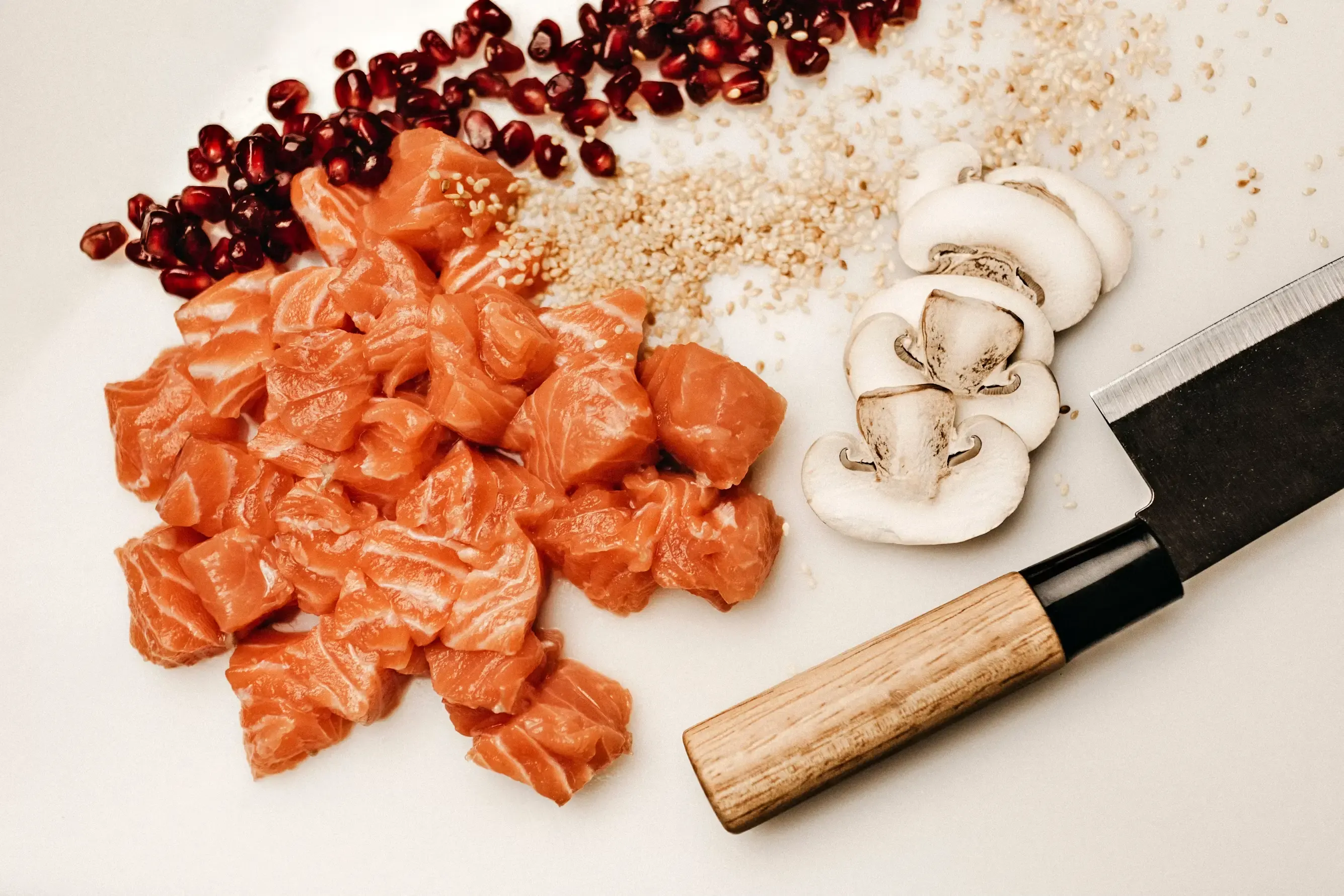 Vista cenital de una superficie blanca con salmón crudo cortado en dados, tres láminas de champiñón blanco, semillas de sésamo esparcidas y granos de granada en la esquina superior izquierda. A la derecha, un cuchillo japonés de hoja oscura y mango de madera clara.