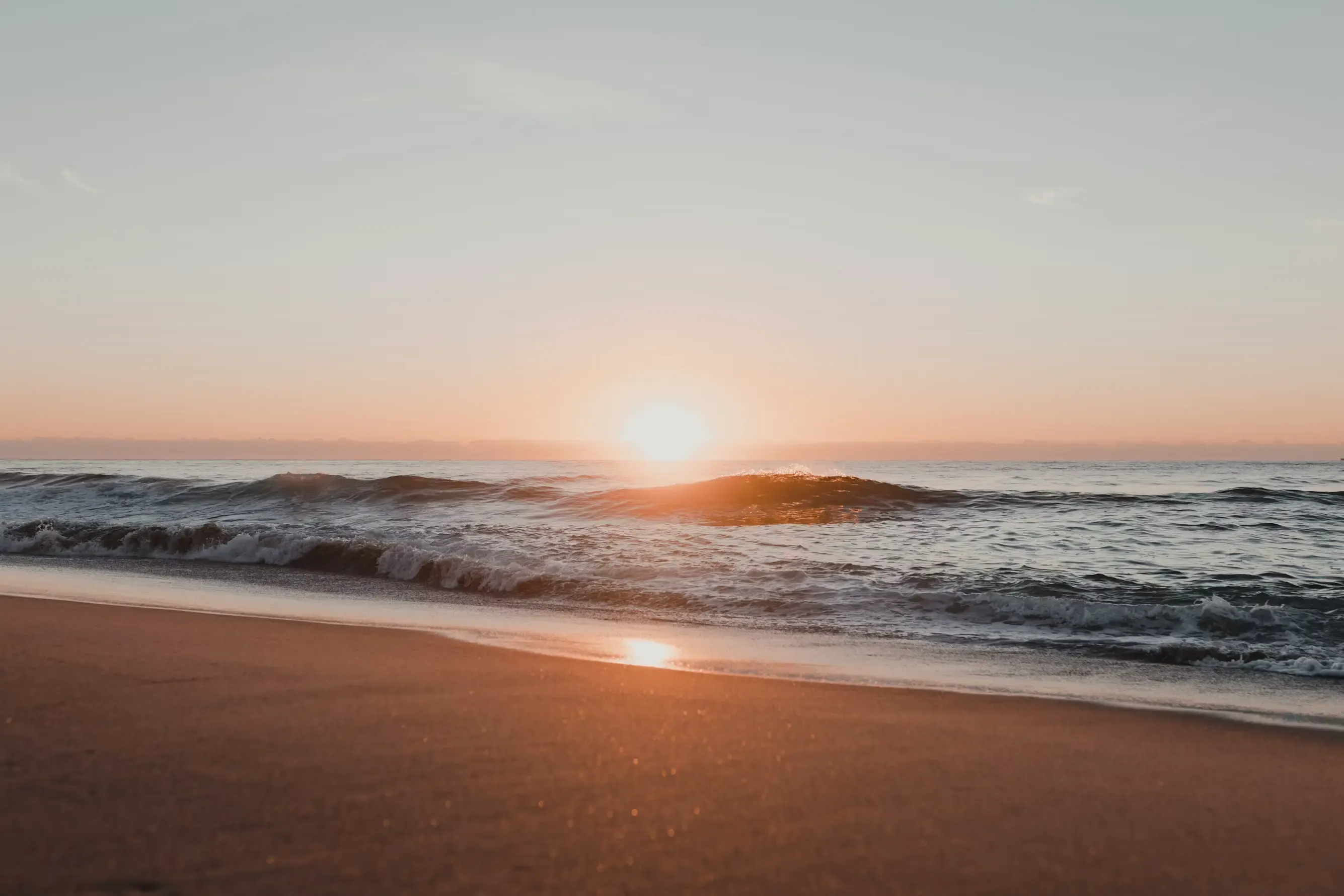 Il sole che spunta all'orizzonte sul mare, con un'onda che si infrange sulla riva e la sua luce riflessa sulla sabbia bagnata della spiaggia.