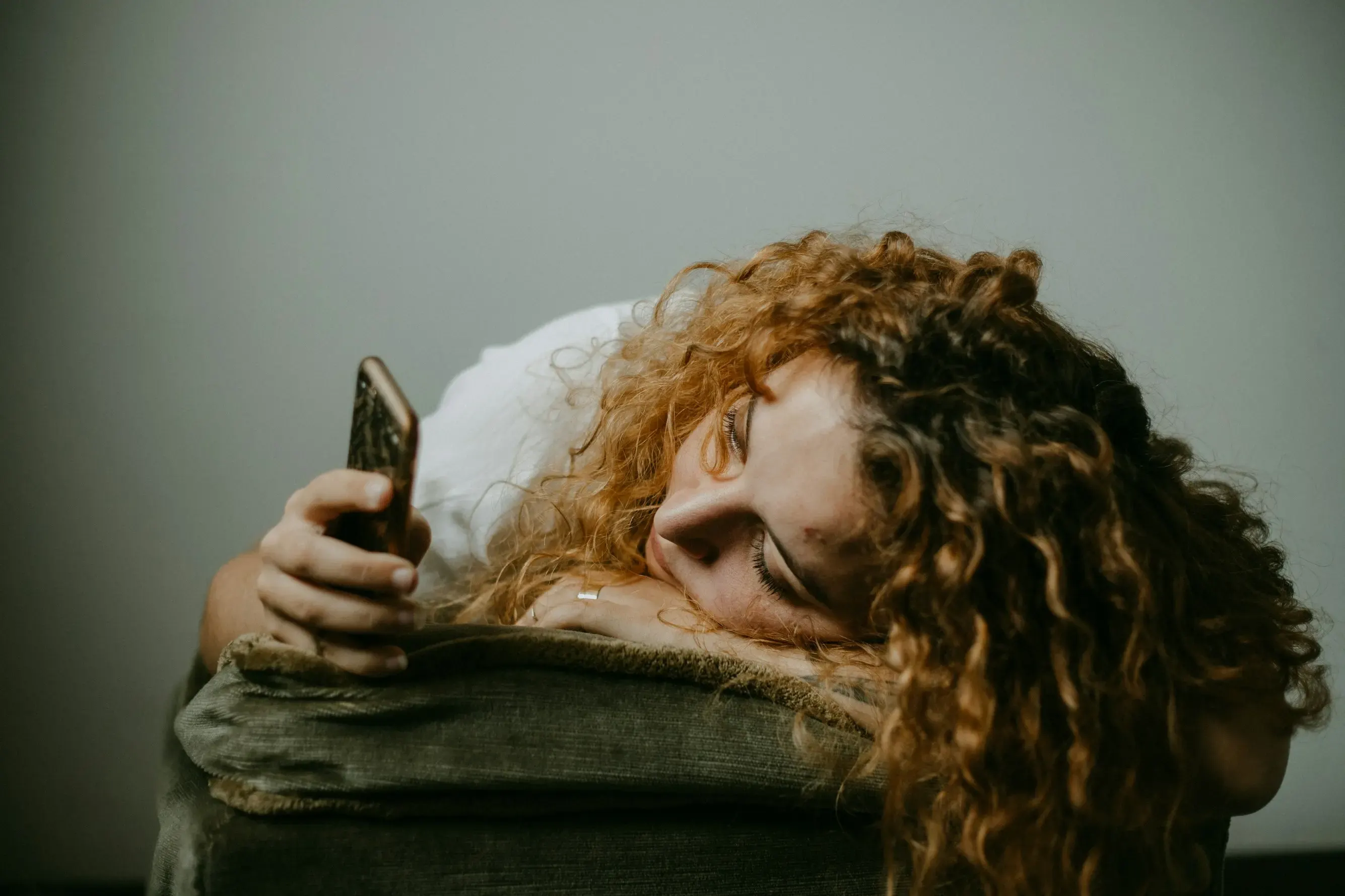 Woman with curly hair lying over the arm of a sofa, absorbed in her phone screen.