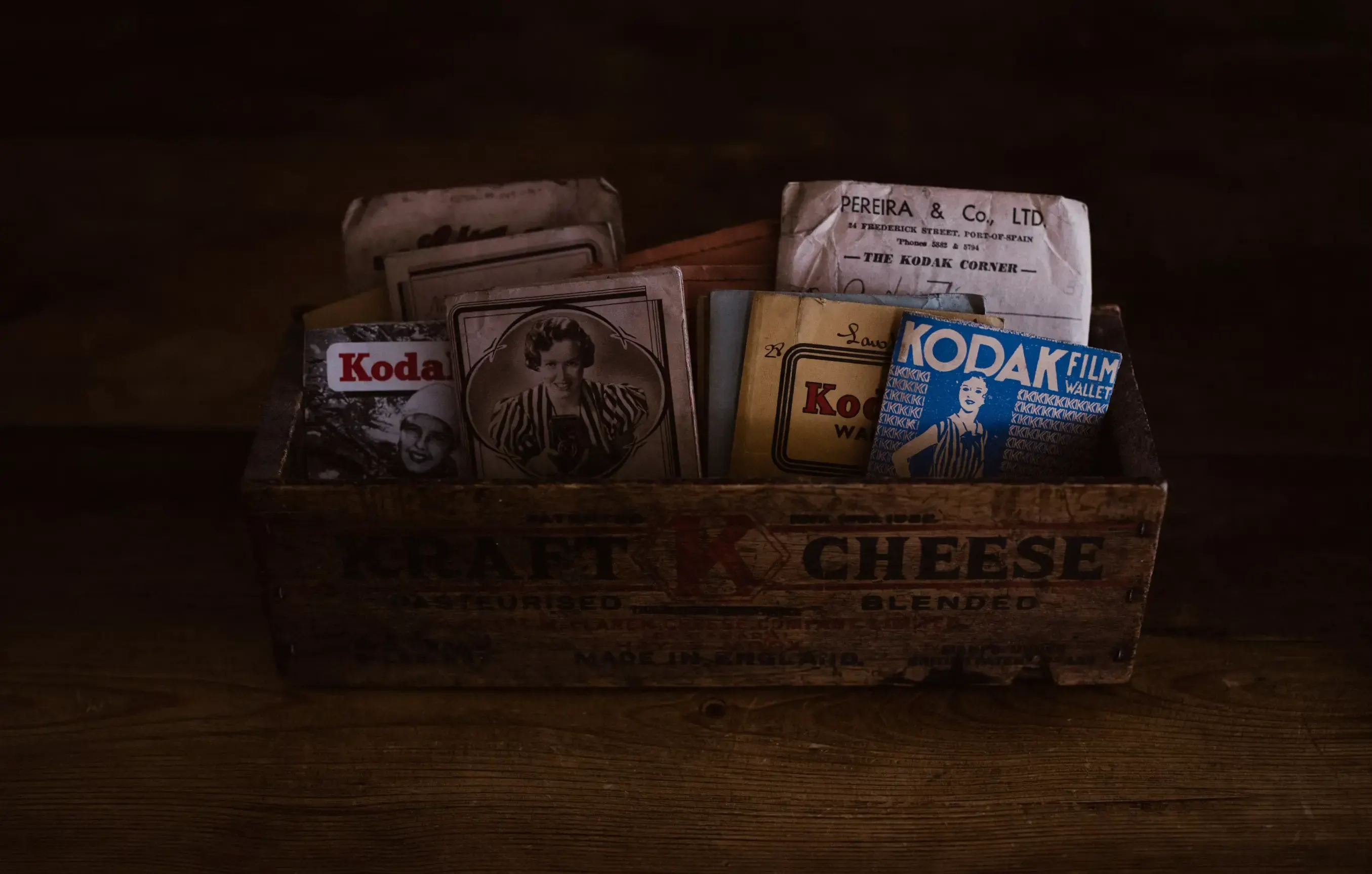 Caja de madera con la inscripción «Kraft Cheese» llena de sobres de fotografías Kodak y retratos antiguos, sobre una mesa de madera en penumbra.